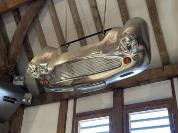 A metal car front, complete with headlights and grille, is mounted as a decorative piece on a wooden ceiling at the Aston Martin Heritage Trust Museum.