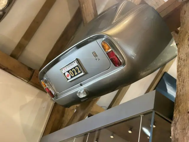 A vintage silver Aston Martin hangs upside down from a wooden structure, with a license plate bearing the number 022, above a glass display case at the Heritage Trust Museum.