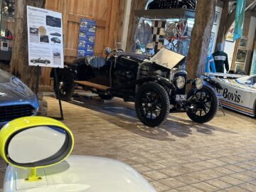 Een vintage Aston Martin met open motorkap wordt tentoongesteld in een museum, samengesteld door de Heritage Trust, naast andere klassieke auto's en informatieve posters. De houten vloer en de bakstenen omgeving completeren het tafereel, waarbij ook verschillende auto-onderdelen zichtbaar zijn.