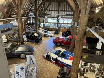 Una exposición interior muestra coches antiguos, incluidos modelos de carreras y clásicos como los de Aston Martin, dispuestos en un desván bien iluminado con vigas de madera y varios paneles informativos y recuerdos del Heritage Trust.