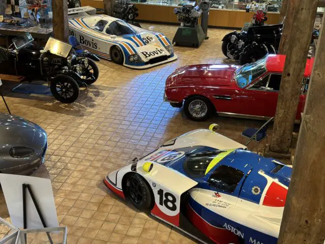 A variety of classic cars and race cars are displayed in a showroom at the Heritage Trust Museum, including an Aston Martin, two Bovis race cars and a red vintage car, surrounded by vintage motorcycles and other automotive memorabilia. This fascinating exhibit offers a stunning photographic record of automotive history.