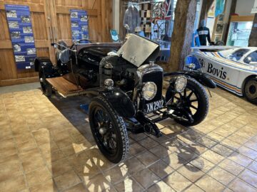 Een zwarte vintage auto met open motorkap wordt tentoongesteld op een tegelvloer in een museumachtige setting, wat de charme van de Aston Martin Heritage Trust oproept. De auto, gedetailleerd in een boeiende fotoreportage, draagt het kenteken "XN 2902.