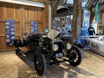 Un Lagonda negro de época con matrícula XN 2902 se expone en el interior de un edificio de madera, que forma parte del Museo Aston Martin Heritage Trust. El entorno, rico en exposiciones de coches históricos y recuerdos, crea un efecto 