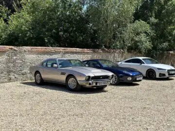 Een zilveren klassieke auto, een blauwe cabriolet en een witte sportwagen staan geparkeerd voor een stenen muur met bomen op de achtergrond. Deze scène, die doet denken aan een tentoonstelling in het Aston Martin Heritage Trust Museum, heeft het wow-effect dat je van zo'n iconische FOTOREPORTAGE zou verwachten.