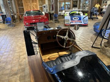 El interior de un coche antiguo con el salpicadero de madera en primer plano, acompañado de un descapotable rojo y un coche de carreras, todo expuesto en el bien iluminado Museo Aston Martin Heritage Trust, crea un innegable efecto sorpresa.