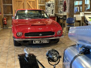 Un coche clásico rojo con matrícula LNV 756L está expuesto en la sala de exposiciones del Museo Heritage Trust, rodeado de vitrinas y puertas de madera al fondo, lo que constituye un fascinante momento de reportaje fotográfico.