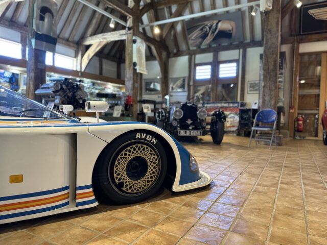 Primer plano de la rueda delantera de un coche de carreras antiguo en el Museo Aston Martin Heritage Trust, con motos clásicas y otro coche antiguo al fondo, todo ello sobre un suelo de baldosas. Las vigas de madera y los detalles del techo contribuyen al efecto sorpresa de esta impresionante sesión fotográfica.