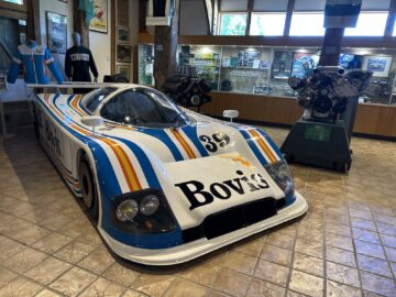 Een tentoonstellingsruimte in het Trust Museum toont een vintage raceauto met de markering "Bovis" in gestreepte kleurstelling, mannequins in racepakken en verschillende autodisplays, waaronder een motormodel.