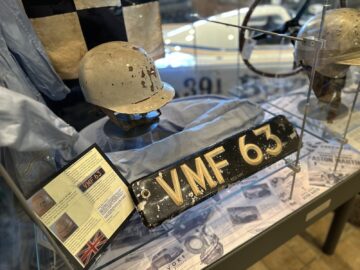 Een vitrine in het Trust Museum toont een versleten helm, het kenteken van een auto met de tekst "VMF 63" en diverse aanverwante items, waaronder beschrijvingskaarten en een achtergrondafbeelding van een Aston Martin Heritage-voertuig met het nummer 391.