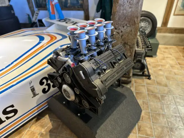 Een close-up van een tentoongestelde automotor, met het opschrift 'Porsche Racing Engine'. De motor is voorzien van meerdere luchtinlaten bovenaan. Op de achtergrond is gedeeltelijk een raceauto met nummer 33 zichtbaar, onderdeel van de tentoonstelling in het Trust Museum met Aston Martin Heritage en exclusieve FOTOREPORTAGE-sessies.