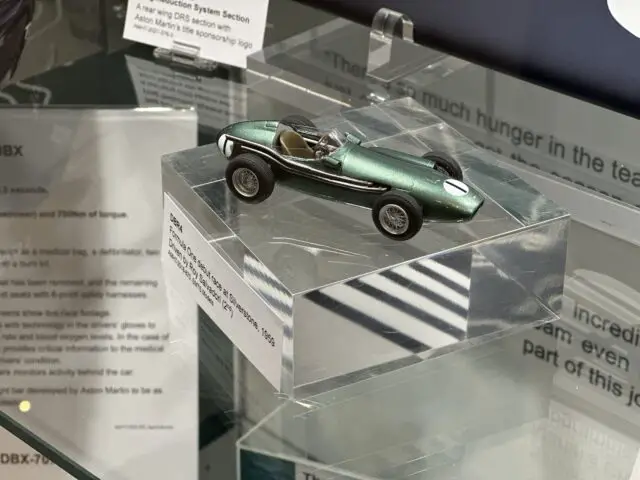 Een kleine groen-zilveren Aston Martin-modelauto uit 1955 wordt in een doorzichtige koffer tentoongesteld in het Heritage Trust Museum, met informatieve tekst op de achtergrond.