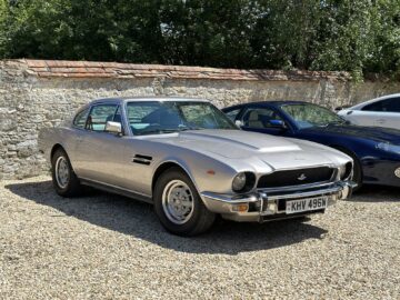 Een klassieke zilveren Aston Martin staat geparkeerd op een grindoppervlak voor een stenen muur met bomen op de achtergrond, alsof hij wacht om tentoongesteld te worden in een FOTOREPORTAGE voor het Heritage Trust Museum.