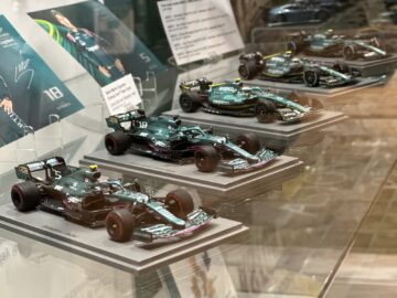 Een vitrine in het Heritage Trust Museum toont vier miniatuur Aston Martin Formule 1-auto's, elk gemonteerd op een basis met daarachter een klein bordje met gedetailleerde specificaties. De groene auto's, versierd met race-emblemen en sponsorlogo's, geven de essentie van Automotive FOTOREPORTAGE weer.