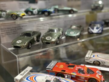 A display case in the museum shows several detailed scale model cars, including green and red racing cars, each marked with different numbers and placed on transparent shelves. It is a fascinating PHOTORY about automotive history, compiled by the Aston Martin Heritage Trust.