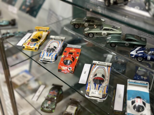 A glass display case at the Heritage Trust Museum shows several detailed scale race cars, including yellow, red and white vehicles of various racing colors, arranged in a neat row on shelves - a perfect opportunity for photo essays.