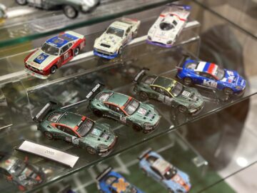 A showcase of model racing cars in racing colors, including several green cars number 58 and Aston Martin number 59 on the bottom shelf, and several other models on the top shelf, reminiscent of a photo essay you might encounter at the Heritage Trust Museum.