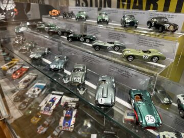 Display case with miniature models of various Aston Martin cars arranged on three shelves. Models include vintage race cars, labeled with information plates. Other car models are visible below, a true photographic record of the Heritage Trust Museum's passion for automotive excellence.