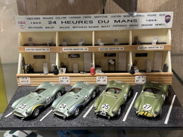 Vier miniatuurauto's van Aston Martin, genummerd 23 tot en met 27, worden tentoongesteld voor een klein model van een pitlane in Le Mans uit 1954. Op de achtergrond staan de namen van verschillende opmerkelijke coureurs, die het rijke Aston Martin-erfgoed vastleggen. Deze prachtige scène hoort thuis in elke fotoreportage van Trust Museum.