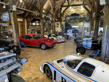 Een ruime kamer met houten balken waarin diverse oldtimers, waaronder een Aston Martin, en automemorabilia worden tentoongesteld. Rondom de auto's zijn verschillende stoelen en informatiestands opgesteld, waardoor een sfeer ontstaat die doet denken aan het Heritage Trust Museum.