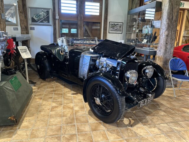 A vintage black Aston Martin with an open hood is displayed at the Heritage Trust Museum, surrounded by automotive memorabilia and exhibits.