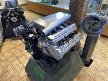 Close-up of a prominently displayed internal combustion engine with visible labels, placed on a stand in a room with additional mechanical exhibits and a tiled floor at the Aston Martin Heritage Trust Museum.