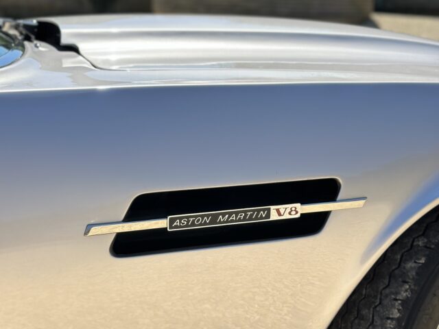 Close-up of a silver Aston Martin V8, with the model's emblem and front wheel, parked outside with a building in the background - perfect for a photo shoot.