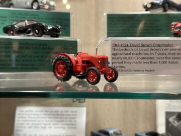 Op een transparante plank wordt een klein model van een rode David Brown Cropmaster-tractor tentoongesteld, vergezeld van een informatief label over de historische productie ervan, waarmee een stukje agrarisch erfgoed in het museum wordt tentoongesteld.