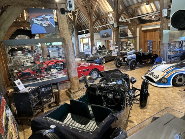 Een goed verlichte tentoonstellingsruimte in het Heritage Trust Museum met verschillende oldtimers, waaronder een zwarte roadster, een rode sportwagen en een elektrisch blauwe Aston Martin-raceauto, omgeven door automemorabilia en kunstwerken. De setting is perfect voor een fotoreportage die de essentie van klassieke auto-elegantie vastlegt.