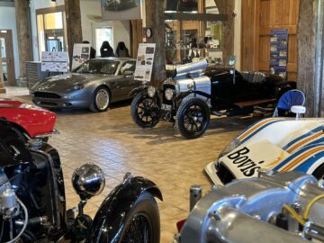 Un efecto sorpresa te espera en el Museo Aston Martin Heritage Trust, donde se expone una colección de coches y motos clásicos y de época en una sala de exposiciones con pilares de madera, carteles informativos y suelo de baldosas. ¡Perfecto para una sesión fotográfica inspiradora!