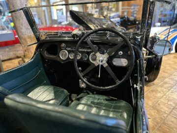 Interior of a vintage Aston Martin convertible with green leather seats and a black dashboard with round gauges, a steering wheel with cross design and a visible rearview mirror, reminiscent of displays at the Heritage Trust Museum.