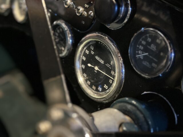 Close-up van het dashboard van een oldtimer met een snelheidsmeter, oliemeter en andere wijzerplaten, waarmee het wow-effect wordt vastgelegd dat doet denken aan een tentoonstelling in het Aston Martin Heritage Trust Museum.