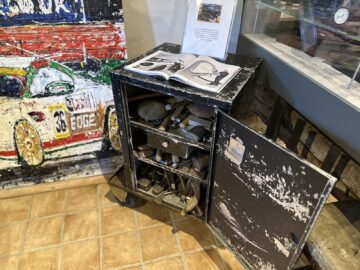 Beneath an open book on machinery is a black, weathered metal cabinet with both a drawer and a door open, showing a variety of old tools. Nearby, a painting of a race car and a glass display case evoke the wow effect reminiscent of a photo report from the Aston Martin Heritage Trust Museum.