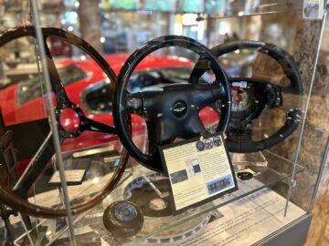 A display case at the Heritage Trust Museum shows several car steering wheels, information boards and an Aston Martin with a red car in the background.