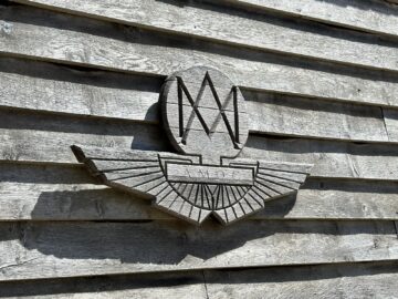 Houten muur met een uitgesneden embleem met "AMOC". Het ontwerp omvat een gestileerde "M" boven een reeks vleugels, waardoor het wow-effect wordt vastgelegd dat hem perfect maakt voor elk Aston Martin Heritage Trust Museum en fotoreportage.