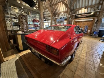 Binnen in het Heritage Museum wordt een rode klassieke auto tentoongesteld, omringd door andere auto-memorabilia en tentoonstellingen. De setting is voorzien van houten balken en een bakstenen tegelvloer, waardoor een perfecte achtergrond ontstaat voor een Aston Martin-fotoreportage.