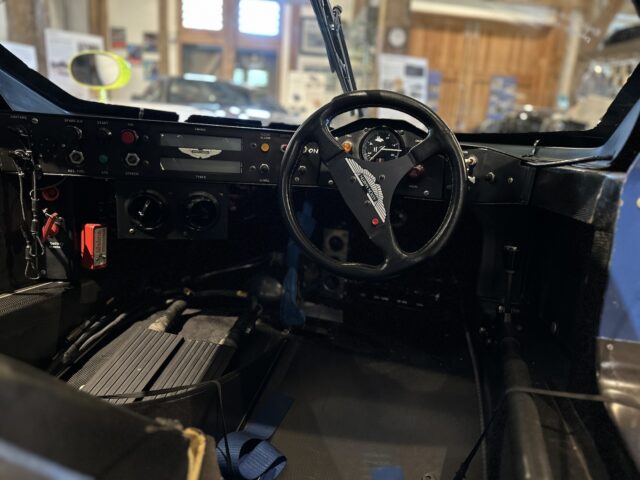 Close-up of a car interior featuring a steering wheel with a logo, controls, pedals and a dashboard with various gauges and switches, reflecting a vintage or race car setup normally found at the Aston Martin Heritage Trust Museum.