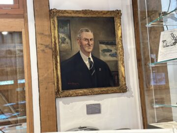 Un retrato enmarcado de un hombre con traje y corbata se exhibe en una pared de madera, flanqueado por estantes de cristal que contienen diversos artefactos y documentos, que recuerdan a una escena de una sesión fotográfica del Museo Aston Martin Heritage Trust.