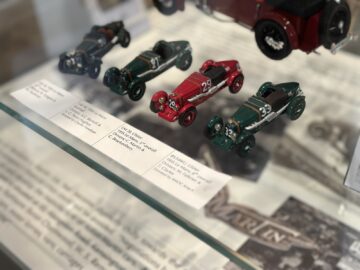 Close-up of vintage race car models displayed on a glass shelf in the Heritage Trust Museum, with informative labels with car specifications and history under each Aston Martin model.