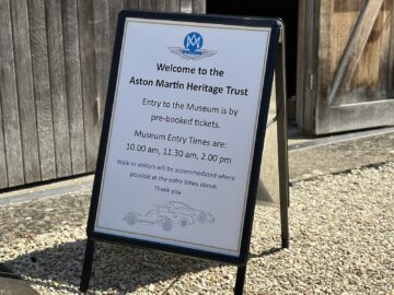 A sign outside the Aston Martin Heritage Trust Museum states that admission is available only with pre-booked tickets, with times at 10 a.m., 11:30 a.m. and 2 p.m. Walk-ins are available based on availability. Capture the wow effect of your visit with a photo report and relive the experience.