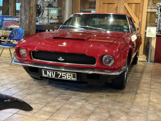 Een strakke rode klassieke auto met het kenteken "LNV 756L" wordt binnenshuis tentoongesteld op een tegelvloer, omringd door automemorabilia in het Heritage Trust Museum. Deze Aston Martin is een hoogtepunt, prachtig vastgelegd in onze laatste fotoreportage.