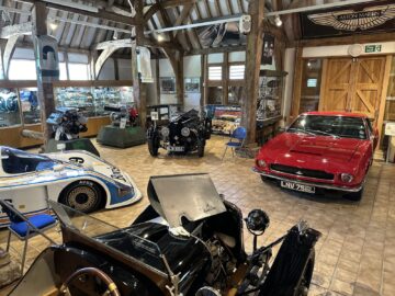 Een kamer met verschillende klassieke auto's, waaronder een rode Aston Martin-sportwagen met kenteken "LNV 755L", een zwarte vintage auto en een witte raceauto. De setting bestaat uit houten balken en automemorabilia, die lijken op een scène uit een uitgebreide fotoreportage in het Heritage Trust Museum.