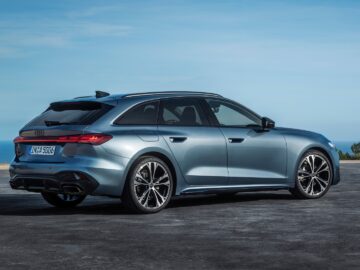 Een metaalgrijze Audi A5 stationwagen staat geparkeerd op een open asfaltoppervlak vlakbij de kust, met de zee en de blauwe lucht op de achtergrond.