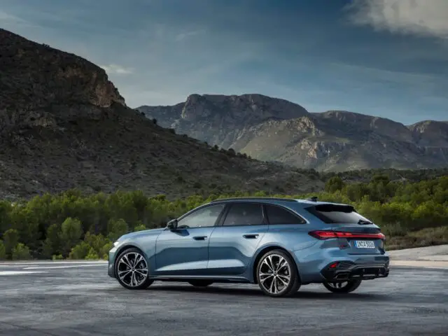 Un elegante Audi A5 sedán plateado está aparcado en un solar asfaltado, con montañas y vegetación de fondo bajo un cielo parcialmente nublado.