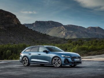 Op een verharde ondergrond staat een Audi A5 blauwe sedan geparkeerd met op de achtergrond een bergachtig landschap. De lucht is gedeeltelijk bewolkt.