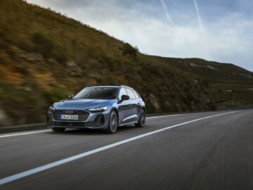 An Audi A5 drives along a winding road with a scenic hill and cloudy sky in the background.