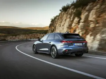 An Audi A5 glides down a winding mountain road with cliffs and greenery in the background under a clear sky.