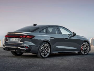 Un Audi A5 sedán gris está aparcado en un claro con el horizonte de una ciudad al fondo. En la matrícula se lee 