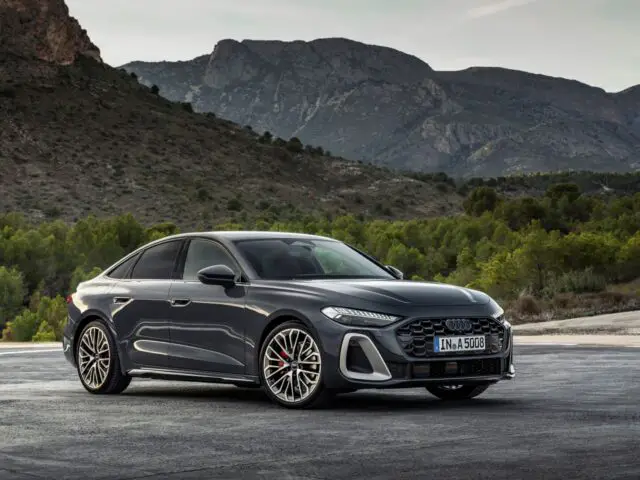 A sleek, gray Audi A5 sedan is parked on an asphalt surface with a background of green trees and rocky mountains under an overcast sky.