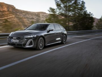 Une élégante berline noire Audi A5 roule sur une autoroute, les mouvements flous indiquant la vitesse. Un paysage montagneux et des arbres sont visibles à l'arrière-plan.