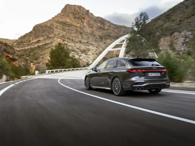 Een strakke zwarte Audi A5 sportback rijdt op een open bergweg met op de achtergrond een witte brug zichtbaar.